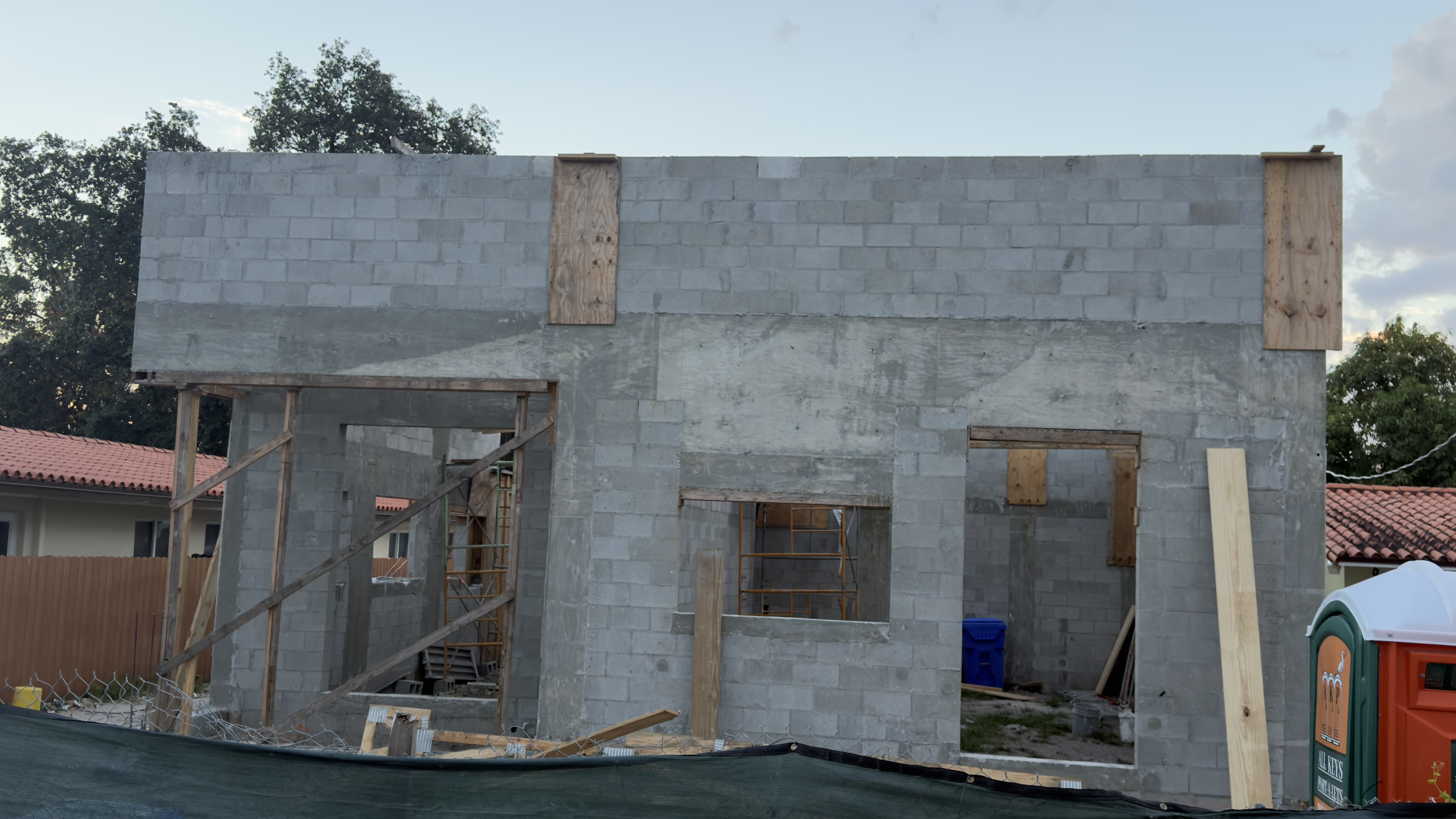 Straight-on view of the block wall from the street, behind chain-link fencing and green privacy screen — the tie beam runs the full width, plywood forms are still strapped at the corners, and three bucked openings frame the scaffolding stacked inside.