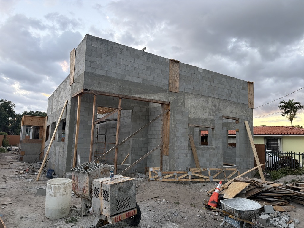 The shell from the opposite corner at dusk, with a mortar mixer and buckets in the foreground, temporary wood truss bracing propped against the wall, and the retained 2017 portion of the house visible at the left.
