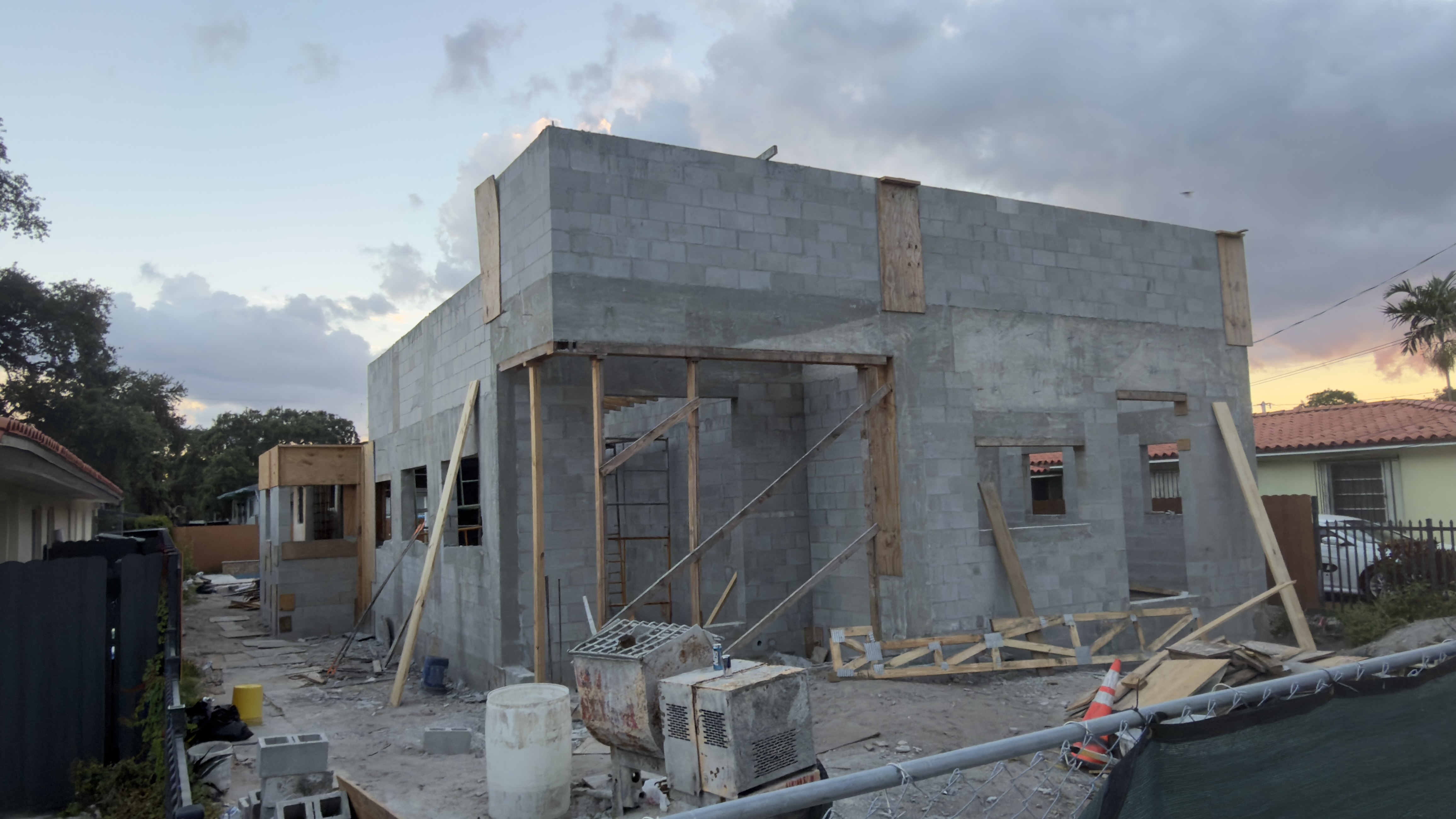 Wide dusk view across the whole site — the full block shell with its tie beam, bucked window and door openings, ladders and shoring inside, stacked lumber and block on the ground, and the neighboring barrel-tile roofs behind.